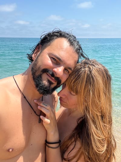 A selfie taken at the beach shows two people, a man and a woman, embracing. The man's face is in the foreground, with his eyes looking at the camera. He has dark, wet hair and a beard. The woman is leaning her head on his shoulder, with her eyes closed and her mouth slightly open. She has long, blonde hair. Both are shirtless and appear to be in the water. The background shows the ocean with clear turquoise water and a blue sky with some clouds. The sun is shining, suggesting it is daytime. The man is wearing a dark necklace with a rectangular pendant. The woman has two bracelets on her wrist. There are tattoos visible on the man's hand, and what appear to be freckles on his chest. The scene evokes a sense of intimacy and romance.