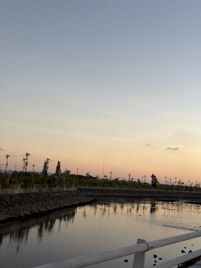 The image depicts a calm body of water reflecting the soft hues of a twilight sky. A stone embankment lines the water's edge, topped with a low fence and then a row of stylized trees or light fixtures resembling birds. In the background, several ornate statues, possibly traditional Indonesian figures, are visible amidst lush greenery. The scene suggests a public park or waterfront area in South Denpasar, Indonesia, at the end of the day.