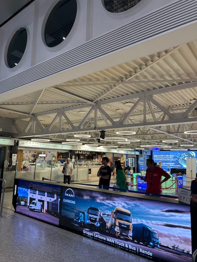 The image captures a scene inside a public space, likely an airport terminal, given the architectural elements such as the elevated walkways, distinctive circular windows, and overhead lighting. In the foreground, a large LED screen displays an advertisement for "Gruppo Carbone MAN Truck & Bus | Rende," featuring images of trucks and vans. Several individuals are visible in the midground, some interacting with the display and others walking past. Behind the LED screen, there are various retail establishments, including a "PAPIILLA food explorers" shop and a "pizzeria," indicating a commercial area. The overall environment appears to be modern and well-lit, with a focus on commercial activity. The text on the screen and the signage for the shops suggest a location in Italy, and specifically the "Rende" mention on the advertisement points towards the area near Cosenza, Calabria, and Lamezia Terme is within the same region.