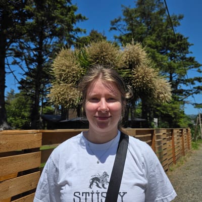 A young woman stands outside in front of a wooden fence and trees. She is wearing a white t-shirt with a black strap over her shoulder. The t-shirt has a graphic of a lion and the word "STUSSY" printed on it. The woman has light skin, light brown hair, and blue eyes. She is smiling and looking at the camera. The background consists of lush green trees and a bright blue sky. The location is Cannon Beach, Oregon, United States of America.