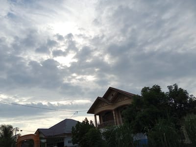 The image depicts a cloudy sky over a residential area in Blangkeude, Indonesia. In the foreground, two houses are visible. The house on the right is a two-story structure with a distinctive roofline and a balcony. The house on the left appears to be a single-story building with a dark-tiled roof. Lush green vegetation, including tall grasses and trees, surrounds the houses. An orange archway with writing, presumably an entrance, is partially visible on the left side of the image. Power lines stretch across the sky, adding to the urban landscape. The overall scene is one of a tranquil residential setting under a dramatic, overcast sky.