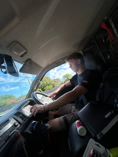 A man with short brown hair is driving a vehicle. He wears a dark t-shirt. His hands are on the steering wheel. The interior of the vehicle is visible, including the dashboard, sun visor, and overhead storage. Through the windshield, a view of trees and a partly cloudy sky is visible. Based on the presence of trees and residential structures and knowing the location, it is likely a street or residential area in Sutherland, Australia. Other items visible include a drink can, a book, and packaged tools.