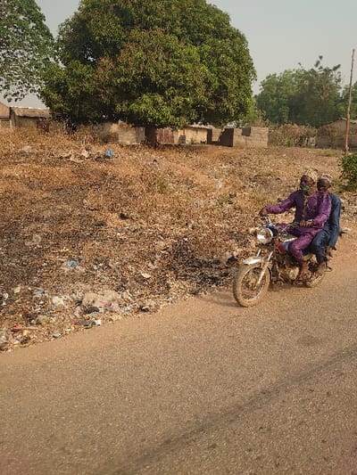 Two adult males are observed on a silver motorcycle traveling on an asphalt road in Osara Gada, Nigeria. The driver, positioned in front, wears a purple long-sleeved garment with a matching patterned cap and a light-colored face mask. The passenger, seated behind the driver, wears a blue long-sleeved garment and a patterned cap similar to the driver's. Both individuals are facing forward.
To the left of the road, a wide, unpaved shoulder is visible, covered predominantly with dry, brown vegetation and scattered refuse, including plastic bags, paper, and other debris. In the mid-ground, a large tree with dense green foliage stands prominently. Behind this tree and extending across the background are several low-lying structures constructed from concrete blocks and corrugated metal, indicating rudimentary buildings or fences. Further back, additional trees with green foliage are present under a clear sky. The ground throughout the background beyond the immediate roadside appears dry and sparsely vegetated.