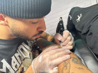 A man in a black beanie and graphic t-shirt, wearing translucent gloves, tattoos a person's arm or leg. The tattoo artist is focused, holding the tattoo machine with both hands as he works. The recipient of the tattoo already has several other tattoos. A portion of the leg is visible, as is an arm of the artist, which also features tattoos. The background shows a partially visible person in dark clothing with a white design and a white brick wall.