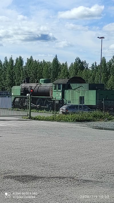 A weathered, green steam locomotive with the number "1011" visible on its side is parked on a railway track in Otanmäki, Finland. The locomotive is partially obscured by a metal fence in the foreground. A dark-colored car is parked behind the fence, near the locomotive. The scene is set against a backdrop of dense green trees and a sky with scattered white clouds. The overall impression is one of a historical or abandoned site.