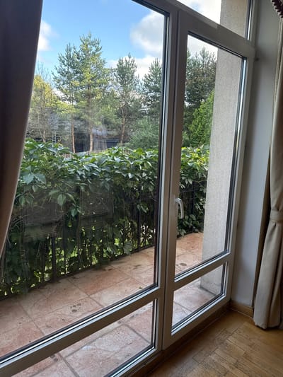 A light-filled scene shows a view through a large, multi-pane sliding glass door. The interior features a wooden floor and a light-colored curtain on the right. The exterior view reveals a balcony with terracotta tiles and a metal railing covered in dense green ivy. Beyond the balcony, several tall pine trees are visible against a blue sky with scattered white clouds. The background shows residential buildings with dark roofing and wooden fences. The location is specified as Yukki, Russia.