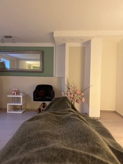 The image displays an interior room in Callbach, Germany, from a low, first-person perspective looking over a person's legs covered by a textured, olive-green blanket. In the mid-ground, to the left, a white two-tiered side table holds several small pink product containers and a glass object, with a magazine visible on the lower shelf. Adjacent to the table is a black velvet armchair with a brown cushion, mounted on a wooden swivel base. Further to the right, partially obscured by the blanket, a floral arrangement with pink blossoms and branches is positioned in front of a light-colored wall. The background on the left features a large rectangular mirror with an ornate silver-colored frame, mounted on a wall painted sage green above a cream-colored panel. The mirror reflects a brightly lit interior space. A ceiling-mounted light fixture with two spotlights is visible above the mirror. The right background consists of plain cream-colored walls extending into a corner, suggesting a hallway or an adjacent room. The ceiling is white, with recessed lighting, and the floor is light-colored wood or laminate. Artificial lighting illuminates the scene. No discernible individuals, specific actions, or interactions are present beyond the covered lower body in the immediate foreground.