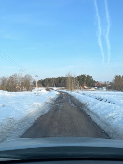 The image depicts a rural or semi-rural winter-to-spring transition scene in Voronino, Russia, viewed from the interior of a vehicle. A narrow, unpaved or heavily worn asphalt road, dark with moisture and bare patches, extends centrally from the foreground into the mid-distance, curving slightly to the left. The road is flanked on both sides by fields covered in snow, which appears partially melted and dirty along the edges of the road.
To the left of the road, utility poles with multiple overhead power lines are visible against the sky, positioned amongst bare deciduous trees and some distant evergreens. Scattered residential structures, partially obscured by trees, are visible in the background on the left.
To the right of the road, fields of snow transition into a dense stand of evergreen trees, predominantly pines, behind which several houses with varying roof colors (red, dark, light) and white fences are identifiable.
The sky is a clear, light blue with two distinct, parallel white contrails trailing horizontally across the upper right quadrant. No individuals or active interactions are visible in the scene. The overall lighting suggests daytime conditions.