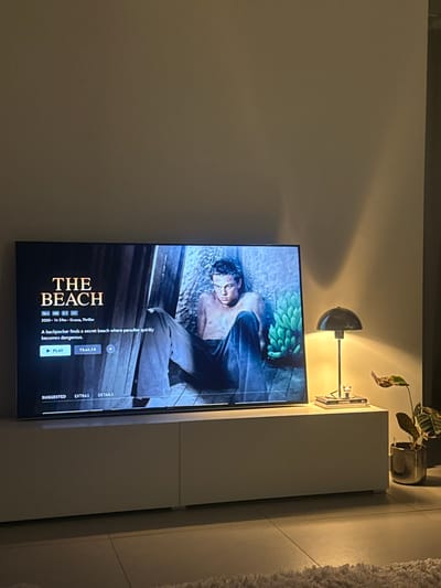The scene depicts an indoor living space located in Tielrode, Belgium. A large, illuminated flat-screen television dominates the center, resting on a low, white, modular TV stand. The television screen displays the title sequence for the film 'THE BEACH,' featuring an image of actor Leonardo DiCaprio, shirtless and wearing light trousers, against a textured background with green bananas. On-screen text includes the title, '16+,' '2000,' '1h 59m,' 'Drama, Thriller,' and a synopsis stating, "A backpacker finds a secret beach where paradise quickly becomes dangerous." Interactive 'PLAY' and 'TRAILER' buttons are visible. To the right of the television, on the TV stand, a lit black mushroom-shaped table lamp with a brass stem casts light and a shadow on the light-colored wall. Beneath the lamp, two stacked books, with 'CHANEL' visible on the top spine, support a small, light-colored decorative figurine. A potted plant with patterned leaves in a reflective gold-colored container stands on the tiled floor to the far right. A portion of a light-colored shaggy rug is visible in the lower right foreground. The room is dimly illuminated, primarily by the television and the table lamp.