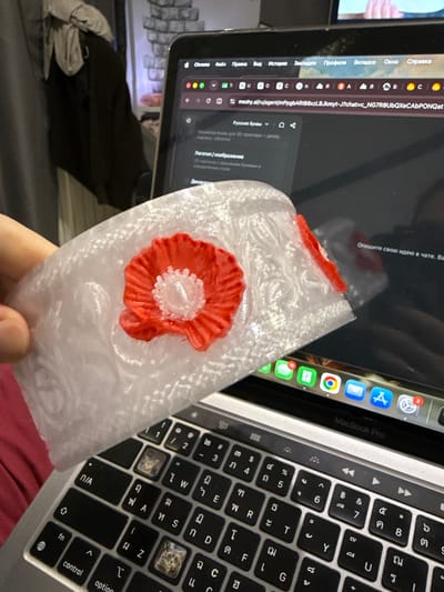 The image shows a close-up of a person's hand holding a white, textured object with a red and white poppy design on it. The object appears to be made of plastic or resin, possibly a decoration or a handcrafted item. In the background, a laptop screen is visible, displaying a web browser with several tabs open. One tab shows a webpage with Russian text, including "Chrome," "Файл," "Правка," "Вид," "История," "Закладки," "Профили," "Вкладка," "Окно," and "Справка." Another tab has a URL that appears to be for a website called "meshy.al." The laptop keyboard is also partially visible, with some keys showing Thai characters and others with decorative stickers of cats. The overall setting appears to be indoors, likely a home or office, with the lighting suggesting it's daytime. There are no clear indicators of the time of day or weather conditions. The focus is on the poppy object and the computer screen, suggesting the person might be using the computer to design or order such items, or perhaps researching crafts. The presence of Russian text and a laptop with a Thai-language keyboard hints at a diverse digital environment.