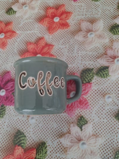 A close-up, overhead shot features a grey ceramic mug with the word "coffee" embossed on its side. The mug is positioned on a white crocheted surface adorned with colorful embroidered flowers in shades of pink, coral, and light peach, along with green embroidered leaves. The scene is illuminated by natural light, casting subtle shadows. The overall aesthetic is domestic and decorative. The image is likely from Fălticeni, Romania, given its domestic subject matter and common decorative styles found in the region.