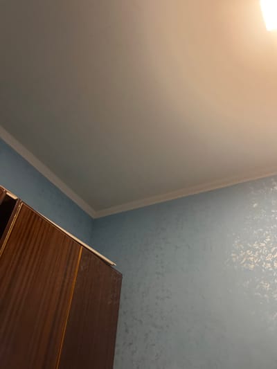 The image shows an interior view of a room, likely a bedroom, with a light blue textured wallpaper. A portion of a dark brown wooden wardrobe is visible on the left side of the frame, with a light-colored edge on top. The ceiling is off-white and there is white crown molding where the walls meet the ceiling. The lighting suggests it might be nighttime or that the room is dimly lit, with a bright light source, possibly a lamp, illuminating the upper right portion of the ceiling. There are no people or animals present in the image, and no specific actions are taking place. The scene depicts a static interior environment. The location in Jrarbi, Armenia is not visually discernible from the provided image.