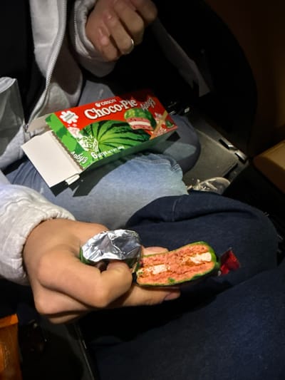 A person is seated, presumably in a car, holding a cross-section of a watermelon-flavored "Choco Pie" snack manufactured by Orion. The snack has a bright green exterior and a layered pink and white interior. The person is wearing light-colored casual clothing, including a zip-up sweatshirt. The box for the Choco Pies is visible, indicating it contains 6 individual pies with a net weight of 168g. The scene does not obviously indicate any location in the city of Tsakhkadzor, Armenia.