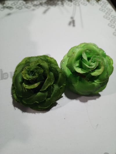Two artificial green roses sit on a white surface. The rose on the left has a deeper, darker green color with a slightly fuzzy texture. The rose on the right is a lighter, brighter green, with a smoother appearance and a subtle glittery sheen. The roses are close together, casting shadows. Fine, stray hairs are visible on the white surface. The background includes faint patterns and sewing pins.