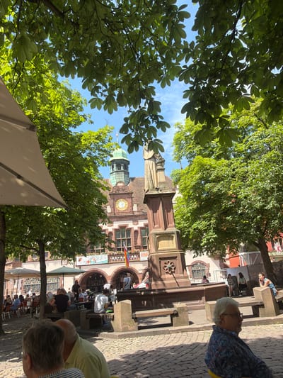 The image depicts a sunny outdoor scene in Freiburg im Breisgau, Germany, likely in a public square. In the foreground, two individuals are partially visible from behind, seated at tables that are not fully in frame. A man with glasses and a patterned shirt is seated on a yellow chair, looking to the right. Behind him, another person with graying hair and a light green shirt is visible from the shoulders up.
In the midground, a stone monument with a statue stands prominently. Behind the monument is a building with an ornate facade, featuring a clock face with Roman numerals and arched windows. A green domed tower is visible on top of the building. Several people are seated at tables under umbrellas and trees, some appearing to be patrons of an outdoor cafe. The ground is paved with cobblestones. Benches are placed around the monument.
In the background, more buildings and trees are visible. The sky is blue with some scattered clouds. The scene appears to be a lively public space, likely a market square or a pedestrianized area. The overall atmosphere is one of a pleasant, sunny day in a European city.