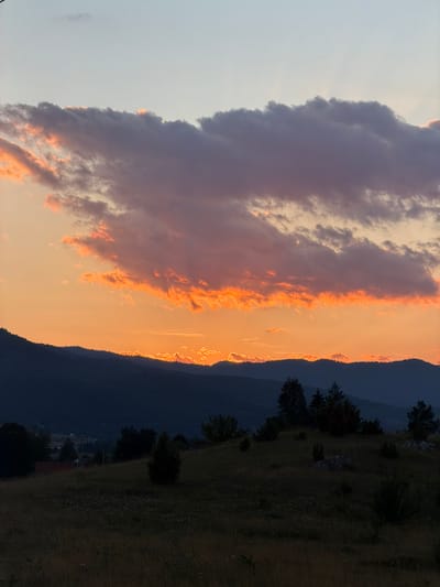 This is a vertical, eye-level shot of a mountainous landscape during sunset. The sky is a gradient of pale blue at the top, transitioning to a vibrant orange, then a soft yellow, before meeting the dark blue silhouette of the mountains. Large, textured clouds dominate the upper half of the image, catching the light of the setting sun and glowing with shades of orange and pink. The sun itself is not directly visible but is suggested by the intense orange light peeking through the gaps in the mountain range.
The foreground consists of a gently sloping, grassy hill dotted with scattered trees and rocks. The grass is a muted brown and yellow, indicative of late summer or early autumn. In the mid-ground, nestled on the lower slopes of the mountains, are a few small, dark buildings with red roofs, suggesting a rural settlement. The overall impression is one of natural beauty and tranquility, with the warm hues of the sunset creating a dramatic and peaceful atmosphere. The location is specified as Donja Brezna, Montenegro.