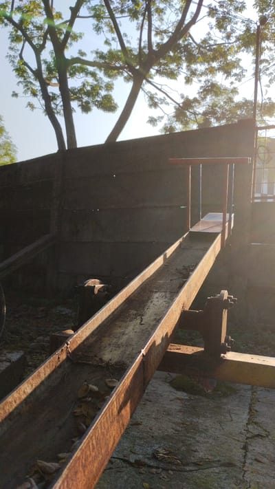 This is a photograph taken outdoors in Trosobo, Indonesia, during the day. The image features a rusted metal seesaw or balance beam, likely part of a playground. The seesaw is positioned diagonally across the frame, with one end elevated and the other closer to the ground. The metal trough of the seesaw appears weathered and contains some fallen leaves. The surrounding area shows a concrete ground with some debris and dried leaves. In the background, there is a dark, textured wall, possibly a fence or building, and above it, the branches of a tree with green leaves are visible against a bright sky, indicating sunlight filtering through. The overall impression is of an outdoor recreational area that may be somewhat aged or disused. There are no people visible in the frame.