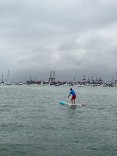 A single person is stand-up paddleboarding on calm water in the foreground, facing away from the camera and towards a busy port in the distance. The person is wearing a blue long-sleeved shirt and red shorts, and is holding a paddle. The paddleboard is turquoise, white, and red. In the mid-ground, numerous sailboats are anchored in a harbor. The background is dominated by a large industrial port with towering cranes, shipping containers, and various vessels. The sky is overcast and gray. The scene is likely the Port of Callao, near La Punta, Peru, a major maritime hub.