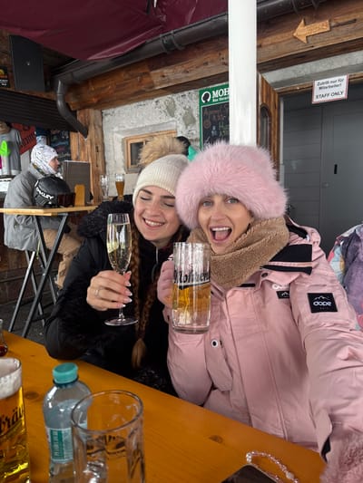 The image shows two women seated at a wooden table within an outdoor or semi-outdoor dining area in St. Christoph, Austria, likely at a ski resort establishment. The woman on the left, with dark hair and wearing a white knit beanie with a beige pompom and a black winter jacket, smiles while holding a flute glass containing a sparkling beverage. The woman on the right, with lighter hair, wears a prominent pink fluffy hat, a brown fleece neck warmer, and a pink winter jacket with a "DOPE" brand patch on the left sleeve. She is holding a large glass beer stein filled with a light-colored beer, her mouth open in an expressive manner.
On the wooden table in the foreground, a plastic water bottle with a blue cap and several empty or partially filled glasses are present, including a tall beer glass with a visible head. In the background, another individual in a grey jacket and a light-colored head covering is seated at a different table to the left. The architectural elements include a rough stone wall, exposed wooden beams, and a dark red awning. Visible signs include a green "MOHREN" logo, a chalkboard menu listing "Aperol 8,50," a white sign on a grey door that reads "Zutritt nur für Mitarbeiter! STAFF ONLY," and a wooden arrow pointing right with "WC / TOILETTEN" indicated.