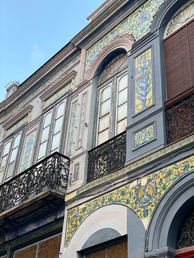 The image depicts an upward-angled view of ornate building facades in Santa Cruz de Tenerife, Spain. The architecture features a mix of grey and pastel-colored facades adorned with yellow and blue tile work depicting animals and floral designs. The building has windows and decorative black wrought iron balconies. The sky is visible in the lower-left corner, indicating the angle of the shot.