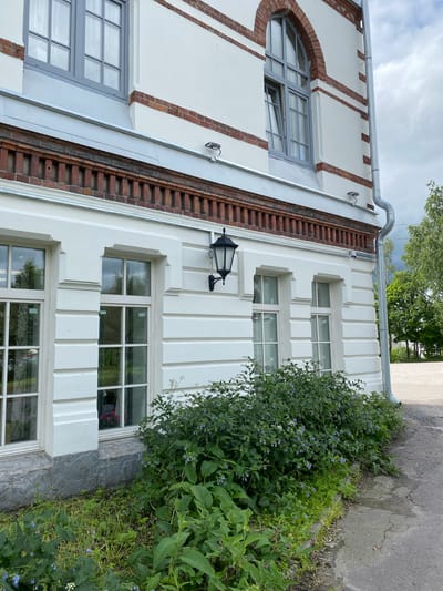 The image shows a close-up view of the exterior of a white building with red brick accents, located in Sortavala, Russia. The building features several large windows with grey frames and a classic black lantern mounted on the wall. Lush green foliage and small blue flowers are growing in front of the building, bordering a paved pathway. A downspout runs vertically along the side of the building. The sky is overcast.