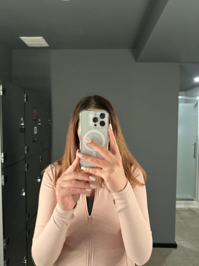 A person is taking a mirror selfie in a locker room. They are wearing a light pink zip-up jacket over a dark top. Their hair is brown and shoulder-length. They are holding a smartphone with a clear case. In the background, there are dark lockers on the left and a grey wall on the right. There is a light fixture on the ceiling. The scene appears to be in a gym or similar facility. There is no information in the image to suggest it is located in Carlow, Ireland.