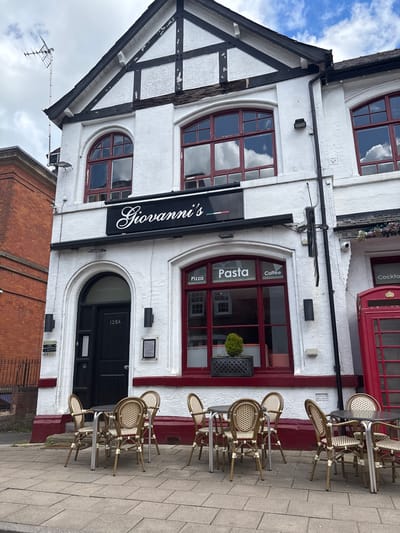 The image shows the exterior of Giovanni's restaurant in Knutsford, United Kingdom. The building is white with black timber framing on the left side, and red trim around the windows and door on the right. A black sign with "Giovanni's" in white script is mounted above the entrance. The arched entrance door is black and labeled "125A". Circular windows above the entrance have red frames and reflect the sky. Words "Pasta", "Pizza", and "Coffee" are printed on the windows, in the same manner as "Cocktails" in a window at the top right of the image. Several rattan chairs and small tables are arranged on the sidewalk in front of the building. A brick building is visible to the left and a wrought iron fence is visible to the right.