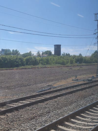 This image depicts a railway scene with several tracks running parallel to each other. The foreground is dominated by the ballast of the railway, with wooden sleepers visible beneath the metal rails. In the middle ground, there is a dense line of green trees, behind which are some buildings, including what appears to be a tall, cylindrical structure resembling a water tower. The sky is a clear blue with a few faint wispy clouds. Overhead, power lines are strung across the frame. The scene suggests an industrial or semi-rural area, likely on the outskirts of a settlement. Based on the provided context, this scene is located in the city of Chulym, Russia. There are no people or animals visible in the image.