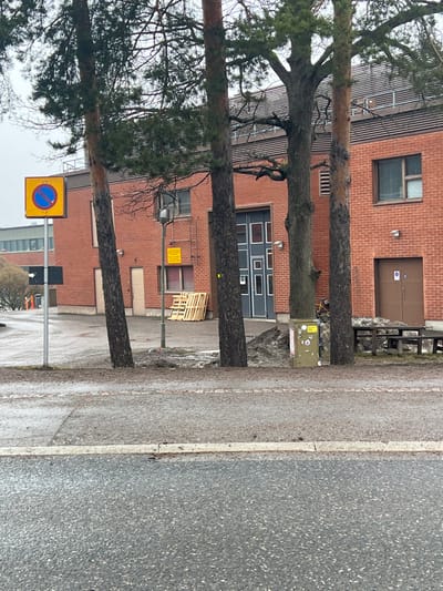 This image depicts an exterior view of a red brick building at Rakentajanaukio 2, within the Aalto University Otaniemi campus area in Espoo, Finland, under overcast daylight conditions. The foreground consists of a wet, dark asphalt road, separated by a curb from a wet, paved pedestrian area. Four mature pine trees with dark trunks and green foliage stand prominently in the mid-ground, partially obscuring the building. Patches of dirty, partially melted snow and mud are visible around the tree bases and near the building.

On the left, a light-grey metal pole supports a yellow square traffic sign with a red circle and blue diagonal line, indicating "No Stopping," with a smaller black sign featuring a white right-pointing arrow below it. Further right, a dark grey street lamp pole holds a smaller, unreadable yellow warning sign. Between the second and third trees from the left, a stack of approximately twelve light-colored wooden pallets rests against the building's facade. A green utility box, adorned with multiple stickers, is situated between the third and fourth trees. A yellow bicycle is partially visible behind this utility box. To the right, a brown wooden picnic table and bench set is positioned on a patch of dirty snow.

The multi-story red brick building features multiple brown-framed windows and doors. A large, segmented dark grey garage door is centrally located. A metal ladder is affixed to the building's wall to the left of this central door. Above the main level, the building incorporates a section with horizontal grey slatted panels. Various outdoor light fixtures are mounted on the facade. No individuals or specific actions are visible within the scene. In the far left background, a lighter-colored building with large windows is partially visible.