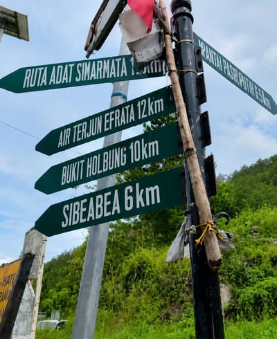 The image shows a directional signpost in a rural or hilly area, likely in or near the city of Sosor Dolok, Indonesia. The signpost is made of dark metal and has several green directional signs pointing left and right, with white text indicating destinations and distances. The signs read: "RUTA ADAT SIMARMATA 1.5km", "AIR TERJUN EFRATA 12km", "BUKIT HOLBUNG 10km", and "SIBEABEA 6km" pointing left, and "PANTAI PASIR PUTIH 6.5km" pointing right. A wooden stick is tied to the signpost with yellow rope. In the background, there is lush green vegetation and a hazy sky. A portion of a yellow sign is visible in the lower left corner, and a white vehicle is partially visible behind the vegetation. The scene appears to be outdoors, during the day, with overcast or partly cloudy conditions.