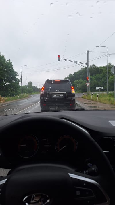 The image is a first-person view from inside a car, looking out through a rain-streaked windshield. The car is stopped behind a black Toyota Land Cruiser Prado on a wet, rural road. The Prado has the license plate "X 657 KM 71". Ahead, a traffic light is red, and a stop sign is visible on the right side of the road. The environment appears to be a wooded area with trees lining the road. The dashboard of the car in the foreground is partially visible, showing the steering wheel and gauges. The sky is overcast and grey, indicating a cloudy or rainy day. The location is identified as Kharino, Russia, by the presence of the Russian license plate format and potentially local road signage, although the sign is partially obscured by glare.