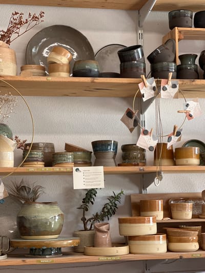 The image displays an interior retail or studio space located in Mafra, Portugal, showcasing a curated collection of handcrafted ceramic pottery arranged on three light-wood shelves against a textured, light-colored wall.
On the top shelf, from left to right, a beige ceramic vase contains dried reddish-brown botanical elements. Behind it, a large, dark grey ceramic plate leans vertically. In front, two stacked, two-toned light brown ceramic cups or bowls with darker bases are present, followed by a squat dark brown bowl featuring a greenish-blue rim. Another dark grey plate is partially visible, behind several dark brown ceramic cups and bowls with glossy dark glazes, some stacked.
The middle shelf features a ceramic pot with dried white flora on the far left. A brass-colored circular frame, adorned with small paper tags secured by mini clothespins, hangs in the foreground, from which a necklace with a ring pendant is suspended. Various ceramic cups and small bowls display diverse glazes, including striped patterns in green, brown, and cream. A white "PRODUCT INFO" card is centrally positioned. To the right, a two-toned cup with a deep blue upper glaze and a light brown base is visible, along with additional brown and dark-glazed ceramic containers, one bearing a subtle "TH" or similar logo.
The bottom shelf on the left supports a large, rounded ceramic pot with a mottled greenish-brown glaze, resting on a circular wooden stand with a turquoise rim. A single feather is placed inside the pot. A white label below the stand reads "SHOPSHELF." To its right, a potted succulent plant in a textured basket is situated next to a small, irregularly shaped pinkish-brown ceramic vase. This vase is placed on a shallow, light brown ceramic tray marked with a logo. Multiple ceramic cups, bowls, and plates in a uniform warm brown and cream color palette are arranged, many featuring the consistent logo. A white label below these items indicates "JEWELRY 625 M35 H30." No individuals are visible, and no dynamic actions or interactions are depicted within the scene.