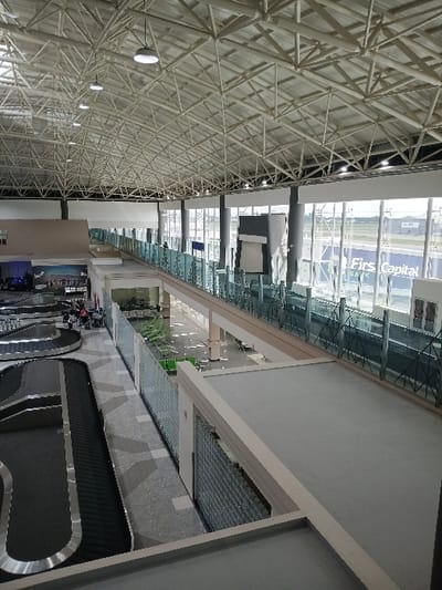 The image depicts the interior of the international terminal at Robert Gabriel Mugabe International Airport in Harare, Zimbabwe, observed from an elevated vantage point.

The scene showcases a spacious, multi-level hall with a high, exposed white metallic truss ceiling structure from which numerous circular pendant lights are suspended. The right side of the hall features extensive floor-to-ceiling glass curtain walls, providing a view of the airport tarmac and a distant "Firs Capital Bank" advertisement visible on an exterior structure.

On the lower left, two large, curved baggage claim carousels, dark gray with silver-colored metal trim, are present. A small group of approximately five individuals is visible near the carousels; one person wears a red top, another a light-colored garment, and they appear to be stationary or moving slowly in the baggage claim area. Adjacent to the baggage claim, separated by a clear glass balustrade with a metal frame, is a public seating area equipped with rows of green-upholstered chairs; two individuals are seated in this area further back.

A light-colored, elevated pedestrian walkway, bordered by clear glass balustrades, traverses the mid-ground. Above this, an enclosed elevated pedestrian bridge, supported by light-colored pillars, runs parallel to the exterior windows. The flooring includes patterned gray carpet in the baggage claim zone and lighter-colored tiled sections in other areas. The overall environment is brightly lit and exhibits minimal pedestrian traffic.