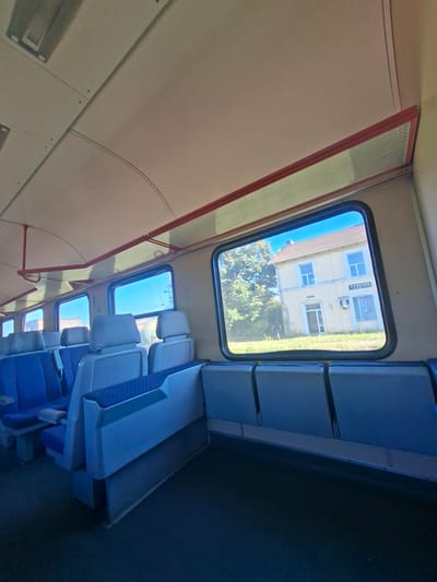 The image shows the interior of a train carriage, with rows of empty blue and gray seats visible. The window of the train offers a view of a yellow building with a sign that reads "КАМЕНЕЦ" in Cyrillic, indicating the location is Kamenets, Bulgaria. The building appears to be a small train station, with trees and a clear blue sky in the background. A person in dark clothing is walking outside the building. The train carriage interior features a light-colored ceiling with exposed conduits and lighting fixtures, and red accent bars along the ceiling.