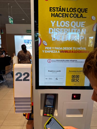 This is a close-up, slightly angled, eye-level shot of a McDonald's self-order kiosk. The kiosk has a large yellow digital screen displaying Spanish text and a QR code for their app. The screen also shows an informational icon and text in Spanish about allergens. Below the screen, there is a payment terminal with a keypad and a card slot. A person's hand is holding a smartphone, the screen of which is visible and appears to be interacting with the kiosk's payment system. To the left of the kiosk is a dispenser for order numbers, with the number "22" clearly visible in large black font on a white background. In the background, a person is visible from the back, walking away from the kiosk, and a woman in a patterned dress is also seen walking away from the camera. The environment appears to be inside a McDonald's restaurant, with other patrons visible in the blurred background. The overall scene suggests a typical fast-food ordering experience with technology. The presence of a "SALIDA" (Exit) sign in Spanish, visible in the upper left crop, suggests the location is in Spain. Given the provided context, the city is identified as Sedaví, Spain.