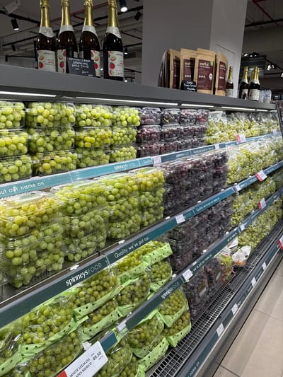 The image shows a refrigerated display case in a supermarket, likely in Ras Al-Khaimah, United Arab Emirates, filled with various types of grapes. The display is organized on multiple shelves.
On the top shelf, four bottles of Spinneys Food Sparkling Apple Juice are visible, with a price tag indicating "20.50". Next to these, several bags of "Oatful Protein Granola" in Chocolate flavor are displayed.
The lower shelves are predominantly stocked with grapes in clear plastic containers and green paper bags. There are two main colors of grapes: green and dark red/purple. The green grapes are presented in a mix of plastic clamshell containers and larger green paper bags. The dark red/purple grapes are primarily in plastic clamshell containers.
Prominent signage along the front of the shelves reads "spinneysFOOD Quality we've put our name on" and "put our name on".
A price tag for "WHITE SEEDLESS GRAPES MEXICO" is visible in the bottom left corner, showing a price of "49.50 DHS" and "per £4".
The environment appears to be a well-lit supermarket aisle with tiled flooring. No people are visible in the image. The overall impression is one of organized retail display of fresh produce.