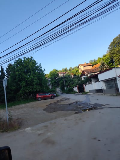 The image depicts a street scene in Rudartsi, Bulgaria, with a clear blue sky overhead. Multiple power lines stretch across the upper portion of the frame, running diagonally from left to right. On the left side of the street, a large, leafy green tree dominates the foreground, with a red SUV parked to its right. Further into the background, houses with terracotta roofs are visible, nestled among more trees on a hillside. The street itself appears to be paved, with a section of it showing puddles of water reflecting the surrounding environment. A signpost is visible near the large tree on the left. The overall impression is a residential area in a somewhat rural or suburban setting.