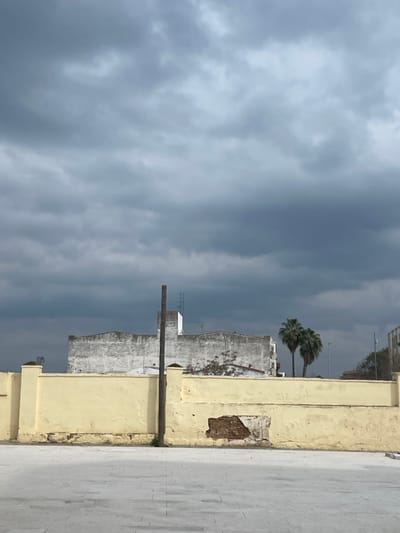The image presents a cityscape scene under a heavily overcast and dark grey sky, filling the upper portion of the frame. Dense, gloomy clouds dominate, suggesting impending rain or a stormy atmosphere.
In the midground, several light-colored, weathered buildings with flat roofs are visible, stretching across the middle of the frame. Rising above these structures are two distinct palm trees, their green fronds providing a natural element against the muted tones of the sky and buildings. A tall, dark pole, possibly wooden or metal, stands vertically in the mid-left, in front of some of these buildings.
To the right, a prominent light yellow or beige wall, likely part of a building or a boundary, runs vertically along the edge. This wall shows signs of wear, with a patch of exposed brickwork or damaged plaster towards its center. To the right of this wall, a paved area, appearing to be light-colored concrete or stone tiles, forms the ground.
No people are visible, and no specific activity is taking place. The time of day appears to be daytime, but the heavy cloud cover gives the scene a somber and dim appearance. The presence of palm trees and the architectural style are consistent with the location of Mérida, Spain. There is no visible text in the image.