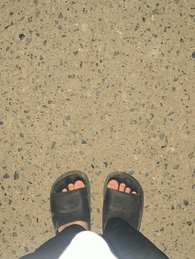 The image displays a top-down view of a person's feet, clad in dark-colored slide sandals, standing on a light brown exposed aggregate concrete surface. The concrete contains numerous small, dark gray, and lighter-colored pebbles. The person's bare toes are visible through the sandal straps. Each sandal has embossed text on its strap, which appears to read "PHOTODESIGN BLABL" (reversed from the viewer's perspective). Dark-colored trousers or leggings are visible around the ankles. A bright white, undefined object or fabric is partially visible at the bottom center of the frame. The scene is located in the city of Manetunong, Indonesia.