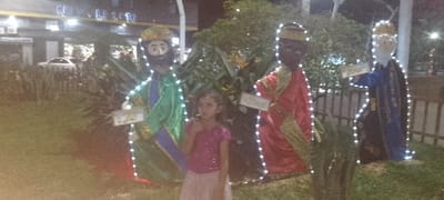 A young girl with a lollipop stands in front of three figures decorated with white lights. The figures, dressed in green, red, and blue robes, appear to represent the Three Kings. A building with lighted signs stands in the background. Trees, grass, and a small wooden fence are visible in the outdoor setting. The scene is captured at night with soft lighting.