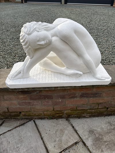 A white sculpture of a nude female figure is lying in a curled position on a white, rectangular base with a textured surface. The sculpture is situated outdoors, next to a brick wall and a paved area. In the background, there is a gravel surface and a building with a dark window frame. The location is likely within the city of Shelve, United Kingdom, judging from the provided information and associated imagery.