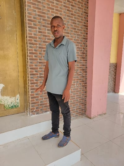 An adult male stands on a white tiled step within a semi-sheltered area in Ebenebe, Nigeria. He wears a slate-grey short-sleeved polo shirt featuring a small emblem on the left chest, black trousers, and blue woven sandals. His left hand is slightly extended forward and open. The background consists of a wall made of reddish-brown bricks with white mortar. To the subject's left, a cream-colored door or panel with some distressed or painted sections is visible. To the right, a structural column painted light pink is present. The floor and steps are composed of white square tiles with dark grout lines.