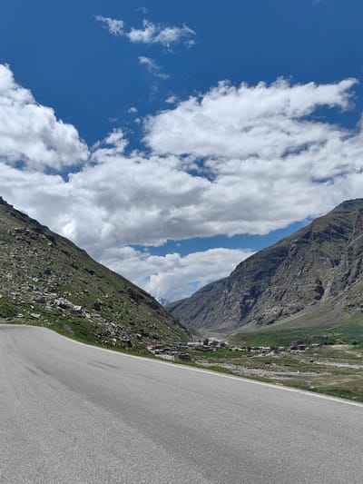 This is a photograph of a scenic mountain pass with a winding road in the foreground. The road curves to the left, leading into a valley. On either side of the valley are steep, rocky mountains covered in patches of green grass and vegetation. The sky above is a vibrant blue, dotted with fluffy white clouds. In the distance, nestled within the valley, is a small settlement with buildings and what appear to be agricultural fields. The image captures a sense of vastness and natural beauty. The location is identified as Kokshar, India.
