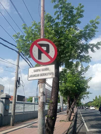 The image depicts a street scene in Surabaya, East Java, under a bright, cloudy sky. A traffic sign prohibiting parking is prominently displayed on a pole. Below it, a white sign reads "SAMPAI RAMBU LARANGAN BERIKUTNYA" which translates to "Until the next prohibition sign." This suggests that the parking restriction applies up to a certain point or until another sign is encountered.

The street is lined with trees, casting shadows on the sidewalk. The sidewalk is paved with red bricks. A metal fence runs along the side of the road, enclosing a building with a grey facade and a blue circular emblem. Vehicles, including cars and motorcycles, are visible on the road, indicating traffic. The overall environment suggests a typical urban setting in Surabaya.