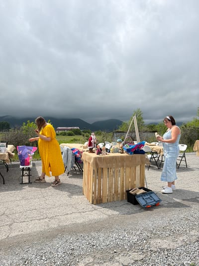 This image depicts an outdoor market or yard sale in Теплоозёрск, Russia, under a cloudy sky. Two women are present. The woman on the left, wearing a yellow polka-dot dress and sneakers, is reaching into a colorful bag on a folding table. She has brown hair. The woman on the right, in a light blue denim dress and white sneakers, is holding a small object and looking down at it. She has dark hair and wears glasses and a white headband. Several tables draped with burlap are set up, displaying various items including a Santa Claus figurine, dolls, bags, and a wooden crate containing a toolbox and other objects. In the background, mountains are visible, with some buildings and trees in the middle ground. The ground is paved with asphalt and gravel.