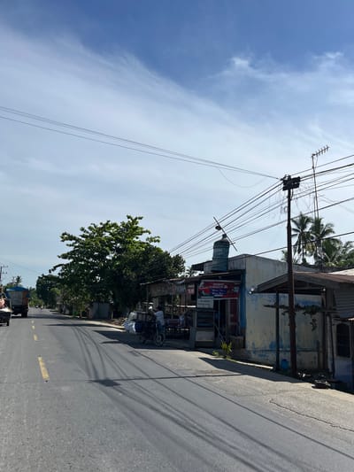 This media file depicts a street scene in Bukitrata, Indonesia. The image is oriented vertically, showing a road on the right side of the frame with a sidewalk and buildings on the left. A prominent feature is the dense network of overhead electrical and telephone wires strung between utility poles. The sky is a bright, clear blue with some wispy white clouds, suggesting it is daytime and the weather is fair.

On the left side of the road, there are several structures, including a building with faded blue and yellow paint, a corrugated metal roof, and a large blue water tank. Various signs are visible, with text that is partially obscured or difficult to read. One sign appears to be an advertisement. A large, leafy green tree grows alongside the road, casting a shadow.

Several people are present, some near the buildings and others on the road. A person is seated on a motorcycle with a sidecar, which is loaded with white sacks. Further down the road, other vehicles and people are visible, suggesting general street activity. The overall impression is one of a bustling, everyday scene in a tropical or subtropical location, characterized by the presence of extensive utility infrastructure and the bright, sunny weather. The time of day appears to be late morning or early afternoon due to the strong sunlight and shadows.
