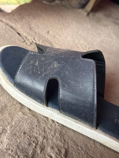 The image shows a close-up, eye-level view of a dark blue, leather-strap sandal resting on a textured, light brown, concrete surface. The sandal's leather strap is slightly worn and dusty, with visible texture and stitching. The sole of the sandal is white and appears to be made of rubber. The background is out of focus, showing a blurred mix of earthy tones and a hint of green. The scene is static, with no people or apparent activity. The setting is an outdoor environment, possibly a pathway or a floor in a city like Bishkek, Kyrgyzstan, given the visual context of the materials and general appearance.