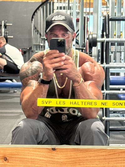 A man with heavily muscled arms and a black cap takes a selfie in a mirror, likely in a gym. He wears a black tank top, gray sweatpants, and gold jewelry (necklace and bracelet). Tattoos are visible on his arm. The mirror has a wooden frame and a yellow sticker with the text "SVP LES HALTERES NE SONT." Gym equipment is visible in the background. The location may be Jemelle, Belgium.