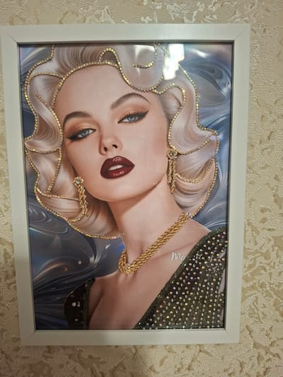 An artwork, likely a framed print with added embellishments, depicts a stylized portrait of a blonde woman with blue eyes, dark lipstick, and elaborate makeup. The artwork is framed in white and hangs on a textured, light-colored wall, possibly with a lace-like pattern. Rhinestones and gold chain elements are applied to the image, accentuating the woman's hair, earrings, necklace, and the shoulder of her dress. The woman's dress appears to be dark and adorned with scattered rhinestones. The background of the artwork is abstract, with swirling blues and grays. The overall impression is one of glamour and opulence. The image is a photograph taken indoors, likely in a residential setting, and there is no discernible connection to Jitiqara, Kazakhstan.