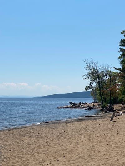This media file captures a serene lakeside scene on a clear, sunny day, likely in the afternoon, given the bright lighting. The image is oriented vertically, presenting a panoramic view of a sandy beach leading to calm, blue water. Scattered driftwood and logs lie along the shoreline, adding a natural, rustic element to the scene.
In the background, a lush, tree-covered hillside rises from the opposite side of the lake, disappearing into a hazy, distant horizon. A few scattered white clouds drift lazily across the bright blue sky, indicating pleasant weather.
The foreground is dominated by the texture of the sandy beach, speckled with small debris. The water is rippled, reflecting the sunlight, and a gentle wave laps at the shore. Along the edge of the water, several small groups of people can be seen enjoying the day, though their specific activities are not discernible. Their presence suggests a leisurely outing, perhaps swimming, sunbathing, or simply relaxing by the water. The overall mood of the image is peaceful and tranquil, evoking a sense of summer relaxation. No visible text is present in the media file.